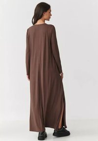 Długa, brązowa, dopasowana sukienka wykonana z miękkiego materiału, posiadająca długie rękawy i boczne rozcięcia, zestawiona z czarnymi butami. Czysty design i minimalne detale.