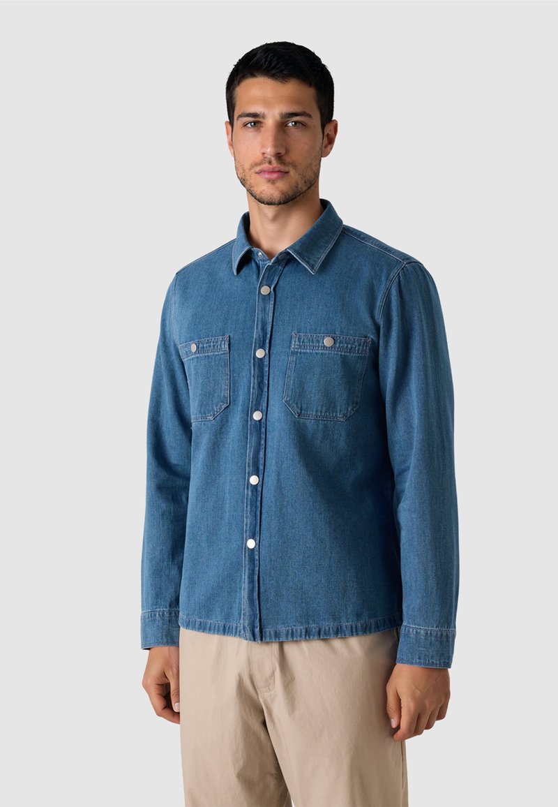 Mann trägt ein blaues Jeanshemd mit zwei Brusttaschen und beige Hosen, steht vor einem einfachen hellen Hintergrund.