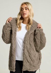 Chaqueta de felpa marrón con cremallera, con un patrón texturizado de punto de cable. Cuenta con una capucha, bolsillos profundos y un corte relajado. Lleva encima una camiseta blanca.