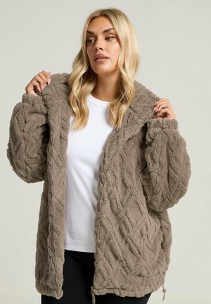 Giacca zip-up fluffy e marrone con un motivo a maglia a treccia. Presenta un cappuccio, tasche profonde e una vestibilità comoda. Indossata sopra una t-shirt bianca.
