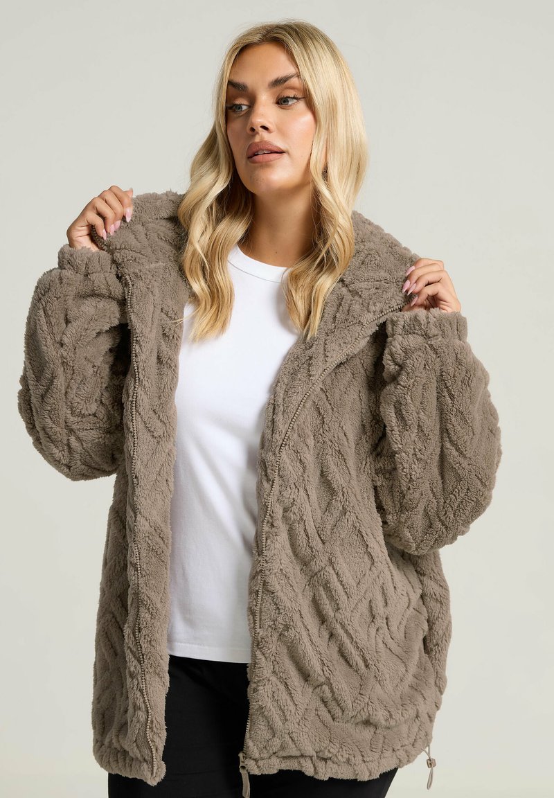 Chaqueta de felpa marrón con cremallera, con un patrón texturizado de punto de cable. Cuenta con una capucha, bolsillos profundos y un corte relajado. Lleva encima una camiseta blanca.