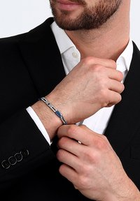 Bracciale in tonalità argento con accenti blu rettangolari, dettagli intricati e un design elegante. Indossato su un polso, in abbinamento a un completo.