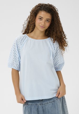 Mladá žena s kučeravými vlasmi, ktorá má na sebe svetlomodrú blúzku s nadýchanými rukávmi s dierkami a rifľovú sukňu na jednoduchom pozadí.