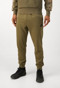 Olīvu zaļi Adidas sporta bikses ar pieguļošu dizainu, elastīgu jostu, sānu kabatām un melnu logo kreisajā augšstilbā. Tekstūrēts audums.