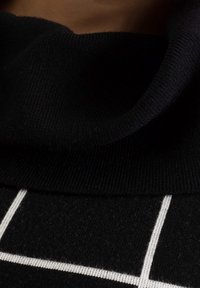 Vêtement tricoté noir avec un col roulé plié, présentant un motif à carreaux blancs, une texture douce et des accents côtelés contrastés au niveau du col.