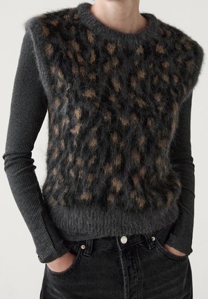 Pull sans manches à imprimé léopard flou noir et marron porté par-dessus un pull à manches longues côtelé gris foncé ajusté et un jean noir.