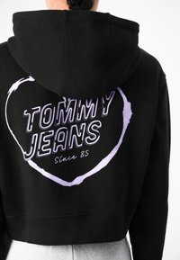 Felpa nera corta con grafica a cuore viola. "TOMMY JEANS" stampato in modo prominente con "Dal 85" sotto, caratterizzata da orlo a costine e cappuccio.