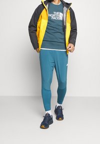 Veste légère jaune et noire sur un sweat à capuche teal avec le logo "THE NORTH FACE", assortie à des leggings teal et des baskets bleu marine.