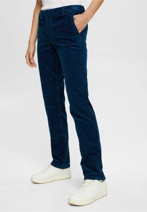 Person iført mørkeblå fløjlsbukser med elastisk talje og hvide sneakers, stående med den ene hånd i lommen mod en ensfarvet baggrund.