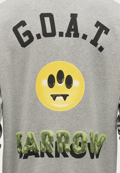 Κείμενο "G.O.A.T.", κίτρινο χαμογελαστό πρόσωπο με τρία μάτια και βαμπιρικά δόντια, και πράσινα χνουδωτά γράμματα που σχηματίζουν τη λέξη "JARROW" σε γκρι ύφασμα.