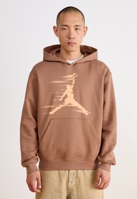 Sweat à capuche marron avec une poche kangourou, présentant un grand graphique beige clair d'une silhouette sautant sur le devant. Texture de tissu doux.