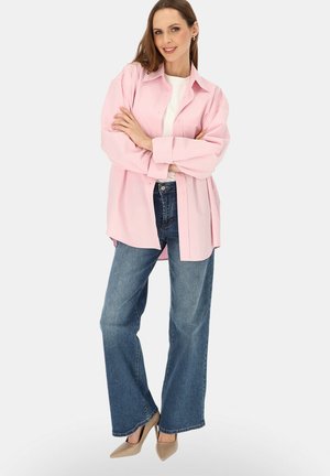 Kobieta stoi z założonymi ramionami, ubrana w oversize'ową różową koszulę, biały top, niebieskie dzwony jeansowe i beżowe szpilki na jednolitym tle.