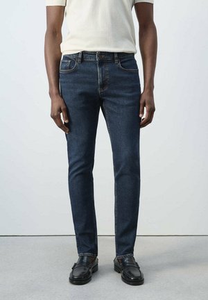 Persona con vaqueros ajustados azul oscuro, mocasines negros y una camisa de manga corta de color claro, de pie sobre un suelo gris.