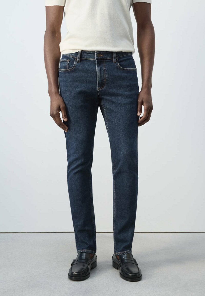 Personne portant un jean slim bleu foncé, des mocassins noirs et une chemise à manches courtes de couleur claire, debout sur un sol gris.