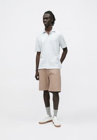 Kortermet, lyseblå teksturert skjorte, kombinert med lysebrune ribbestrikkede shorts. Hvite joggesko og grå sokker fullfører antrekket.