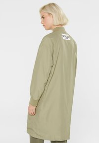 Olivgrüner, langer Bomberjacke mit gerippten Bündchen und Saum, ausgestattet mit einem weißen Etikett mit "SENSES" auf dem Rücken. Glattes, leichtes Material.