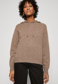 Felpa a maglia marrone, con cappuccio regolabile e polsini a costine. Texture morbida, vestibilità comoda, abbinata a pantaloni neri.