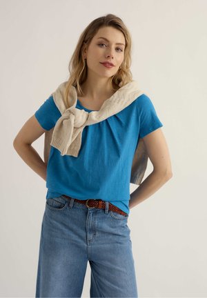 Pruunide juustega naine, kes kannab sinist t-särki ja kreemjat kampsunit üle õlgade, poseerib käed puusas valge tausta ees.