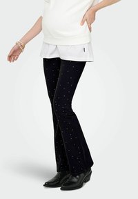 Svarte utsvingte jeans med prikkede detaljer, kombinert med en hvit lagdelt topp; modell iført svarte hælte ankelstøvler.