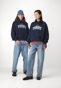 Sudaderas azul oscuro con el logo blanco "TOMMY JEANS", combinadas con jeans azul claro. Los accesorios incluyen una gorra de denim y una camiseta de rayas debajo de la sudadera.