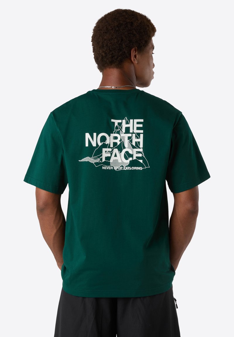 Mand med kort, krøllet hår iført en mørkegrøn T-shirt med hvid "The North Face" tekst og bjergkontur på ryggen, sorte shorts.