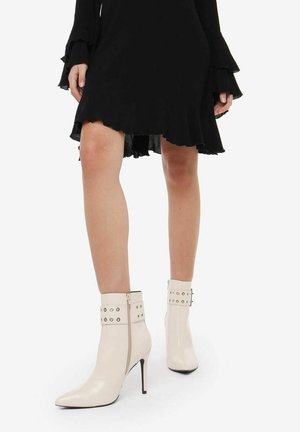 Bottines blanches en cuir synthétique avec un bout pointu et un talon aiguille, comportant des zips latéraux et des accents cloutés sur la sangle de la cheville.