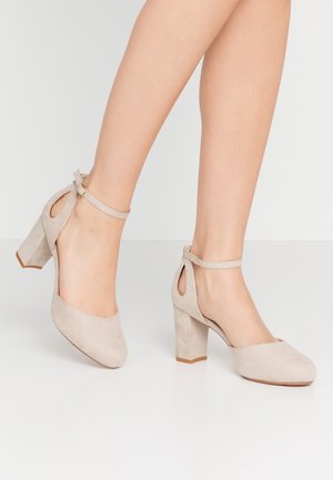 Beżowe zamszowe szpilki z paskiem wokół kostki, kwadratowym noskiem, wycięciami i masywnym obcasem. Gładka faktura i minimalistyczne akcenty.