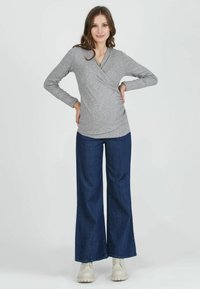 Graues langärmliges Wickeltop mit gerippter Textur, kombiniert mit weit geschnittenen, dunkelblauen Jeans und cremefarbenen Sneakers, vor einem weißen Hintergrund.