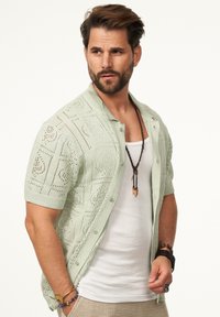 Hellgrüner, gestrickter Cardigan mit kurzen Ärmeln und offenem Muster, Knöpfen vorne und einem Kragen. Tragen über einem weißen Tanktop.