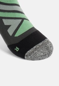 Calzino nero e verde con un design geometrico, dotato di una punta grigia e un motivo testurizzato. Indicatore di misura visibile vicino alla punta.