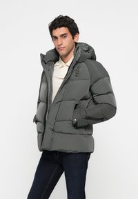 Chaqueta de plumas en gris oscuro, con capucha, bolsillos con cremallera y una textura acolchada audaz. La tela parece ser resistente al agua y duradera.