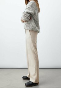 Person standing sideways wearing a speckled beige sweater, cream-colored pants, and black clogs with wooden soles on a gray floor.