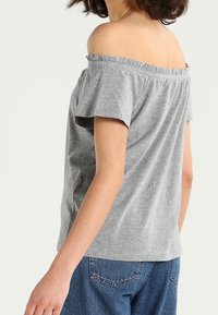 Gray off-the-shoulder top with short sleeves, made of soft fabric. Features a ruffled neckline and a relaxed fit. Paired with denim bottoms.