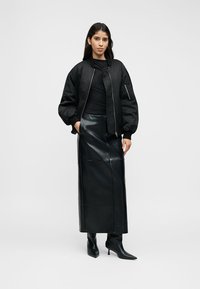 Schwarze Bomberjacke mit gesteppter Textur, kombiniert mit einem langen schwarzen Lederrock. Modell trägt spitze schwarze Stiefeletten. Minimalistisches Design.