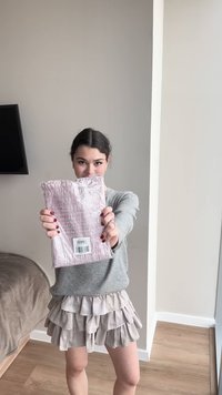 Mujer joven con suéter gris y falda beige sosteniendo un artículo empaquetado en rosa con código de barras en una habitación luminosa con suelo de madera.