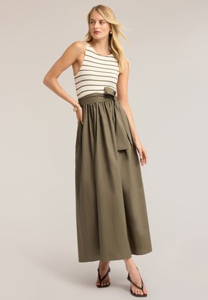 Mujer con una blusa sin mangas a rayas blancas y negras metida en una falda ancha de pierna alta de color verde oliva con un cinturón de lazo y sandalias negras.
