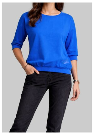 Femme portant un haut à manches longues bleu vif avec les manches retroussées et un jean noir, debout avec une main dans la poche devant un fond uni.