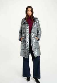Sølvfarvet, puffy yderjakke med hætte, frontlommer og en blank overflade. Bæres over en bordeauxfarvet turtleneck og mørke, vide jeans.