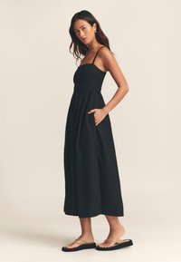 Vestido largo negro con corpiño ajustado y falda amplia, con tirantes finos ajustables y bolsillos laterales. La modelo lleva sandalias planas.