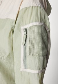 Grøn letvægtsjakke med en kontrasterende lyse beige topsektion. Har en mesh-lomme på armen og synlig lynlås-lukning.