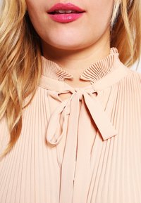 Vaaleanpunainen pleatattu paita, jossa on korkea röyhelökaulus ja solmittava yksityiskohta kaulassa. Sen pinta on sileä ja siinä on pystysuoria poimuja, jotka kulkevat kankaan alas.