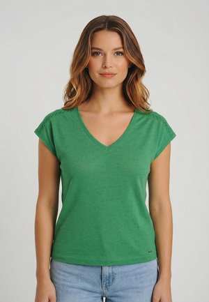 Donna con capelli marroni mossi che indossa una maglietta verde a scollo a V con maniche corte e jeans azzurri, in piedi davanti a uno sfondo chiaro e uniforme.