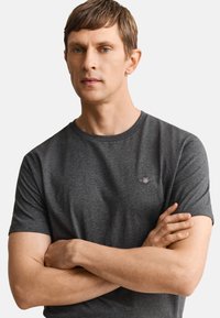 Mann mit kurzen braunen Haaren, der ein dunkelgraues kurzärmeliges T-Shirt trägt und mit verschränkten Armen vor einem einfachen hellen Hintergrund steht.
