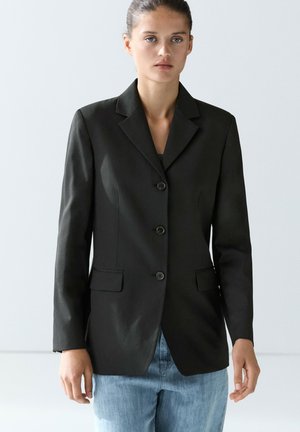 Donna con capelli raccolti indossa una giacca nera abbottonata sopra jeans azzurri, in piedi davanti a uno sfondo grigio chiaro uniforme.