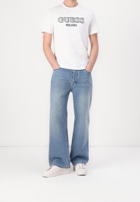 Weißes Baumwoll-T-Shirt mit blauem "GUESS EST. 1981" Grafik, kombiniert mit hellblauen, weiten Denim-Jeans und weißen Sneakers.