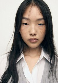 Porträt einer Person mit langen, glatten schwarzen Haaren, die ein weißes Hemd und eine graue, gestreifte Weste trägt, mit auffälligen Sommersprossen auf den Wangen.