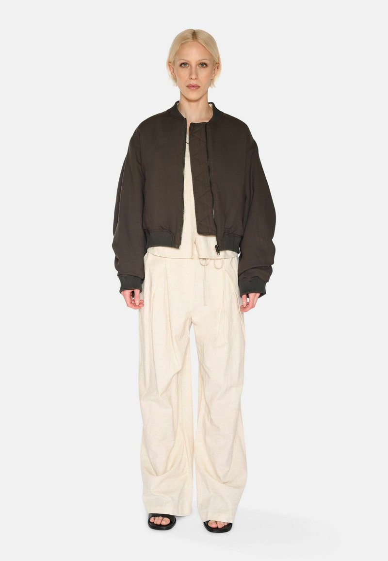 Braune kurze Jacke mit gestepptem Futter und gerippten Bündchen, kombiniert mit lockeren, elfenbeinfarbenen Hosen mit Kordelzug. Vor einem weißen Hintergrund.