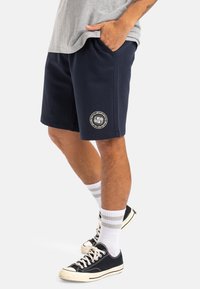 Bermudas azul marino hechas de tela suave, con bolsillos laterales y un parche de logo circular en la pierna izquierda. Llevadas con calcetines grises y zapatillas negras.