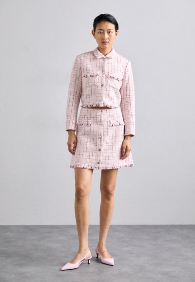 Rosa tweed jakke og skjørt sett med frynse detaljer, fire lommer og sølv hardware. Modellen har på seg rosa spiss tå hæler.
