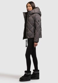 Puffet grå jakke med diamantstepping, en stor hætte og lynlåslommer. Stylet med sorte leggings og blanke sorte støvler.